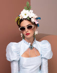 A woman wearing a light blue dress with puffed sleeves, white Frida Portrait by For Art's Sake® sunglasses with ultra-lightweight nylon lenses, and flower earrings poses confidently. She has a floral headpiece adorned with various flowers and leaves atop her dark hair. The background features earthy tones, enhancing her radiant look.