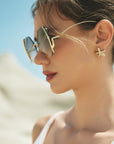 A woman models For Art's Sake® Generation sunglasses with geometric lenses and starfish-shaped earrings outdoors under bright sunlight, set against a blurred sandy background.