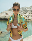 A woman wears For Art's Sake® Generation geometric lenses sunglasses and a white bikini, standing by the water with rocky cliffs, a vibrant leopard print scarf draped over her shoulders.