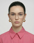 A woman with dark hair in a neat bun wears gold hoop earrings, a buttoned-up pink shirt, and Genius Two unisex opticals by For Art's Sake®, facing forward with a neutral expression against a plain background.