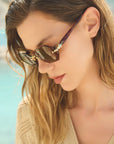 A woman with wavy, light brown hair wears For Art's Sake® Haze geometric sunglasses with rimless thick lenses and a beige top, looking down. The soft blue tones in the blurred background evoke an outdoor, waterside setting.