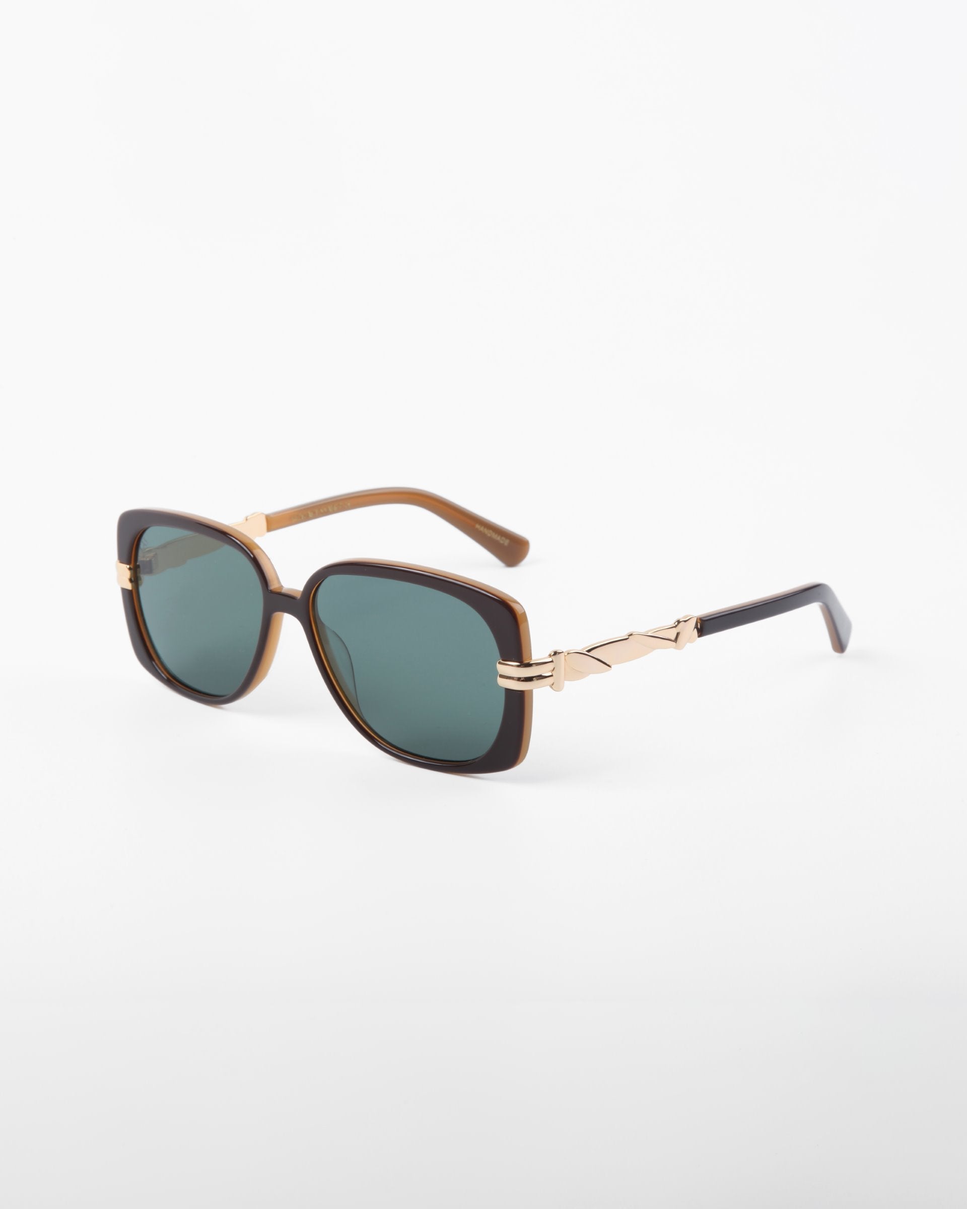 For Art's Sake® Icon-ARC sunglasses feature square green-tinted lenses and handmade acetate with a brown frame and gold accents. These stylish D-frame shades, positioned elegantly on a white surface, offer full UVA & UVB protection.