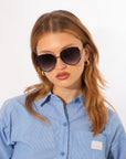 A woman with long, light brown hair models For Art's Sake® Wonderland oversized sunglasses with 100% UV protection, gold hoop earrings, and a blue and white striped button-up shirt against a plain white background.