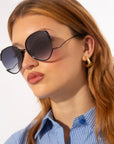 A young woman with light brown hair models For Art's Sake® Wonderland—oversized black round sunglasses with jadestone nose pads and 100% UV protection—paired with gold hoops and a blue striped shirt against a light background.