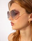 A woman with light brown hair wears For Art's Sake® Wonderland round sunglasses with jadestone nose pads and gold hoop earrings, gazing to the side against a plain light background.