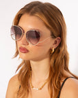 A young woman with light brown hair wears For Art's Sake® Wonderland sunglasses with 100% UV protection, a gold chain necklace with pearls, gold hoop earrings, and a black top, posing against a plain white background.