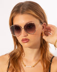 A woman with light brown hair wears For Art's Sake® Wonderland large, round sunglasses with jadestone nose pads, paired with pearl gold jewelry, as she touches her ear and looks to the side against a white background.