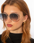 A young woman with light brown hair wears For Art's Sake® Wonderland large, round sunglasses with 100% UV protection, gold hoop earrings, a black top, and a gold chain necklace against a plain white background.