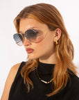 A woman with light brown hair models For Art's Sake® Wonderland sunglasses featuring jadestone nose pads and 100% UV protection, paired with gold hoop earrings and a pearl-accented gold chain necklace, against a plain white background.