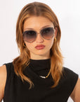 A woman with light brown hair wears For Art's Sake® Wonderland large round sunglasses with 100% UV protection, gold hoops, and a black sleeveless top with a gold chain and pearl necklace, posing against a white background.