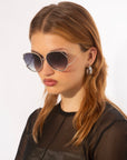 A woman with light brown hair models For Art's Sake® Wonderland sunglasses—large, round frames with jadestone nose pads and 100% UV protection—paired with silver hoop earrings and a sheer black top, against a plain white background.