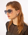 A woman with long, light brown hair wears For Art's Sake® Wonderland round oversized sunglasses and silver hoop earrings. She looks to the side in a sheer black top against a plain white background.