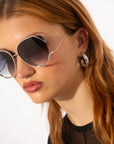 A woman with long, light brown hair wears For Art’s Sake® Wonderland sunglasses with jadestone nose pads and silver hoop earrings. She has a neutral expression, a black top, and the sunglasses provide 100% UV protection.
