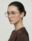 A woman with fair skin and dark hair wears For Art's Sake® Lace square acetate frames with clear jadestone nosepads and a brown top, gazing slightly to the side against a plain light background.