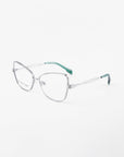 The Lady-S by For Art's Sake® are silver-framed eyeglasses with a subtle cat-eye shape, intricate twisted metalwork, translucent nose pads, and green temple tips against a white background.
