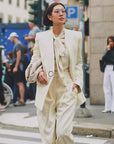 A stylish individual walks confidently down a city street wearing an off-white pantsuit with a matching tie-neck blouse. They carry a bag under one arm and sport UV-protected aviator shades from For Art's Sake® called Explorer Pink. In the background, people are engaged in various activities near a busy intersection.
