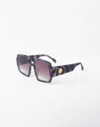 A pair of stylish black and gray marbled sunglasses with large, square, UV-protected lenses featuring a gradient tint. The chunky acetate frame is adorned with a circular gold emblem on each temple, set against a clean white background. These are the Moritz by For Art's Sake®.