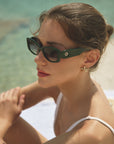 A woman in dark green For Art's Sake® Mene Cat-Eye sunglasses and gold seashell earrings sits by clear blue water, her hair tied back as she gazes off to the side in a white swimsuit, looking relaxed.