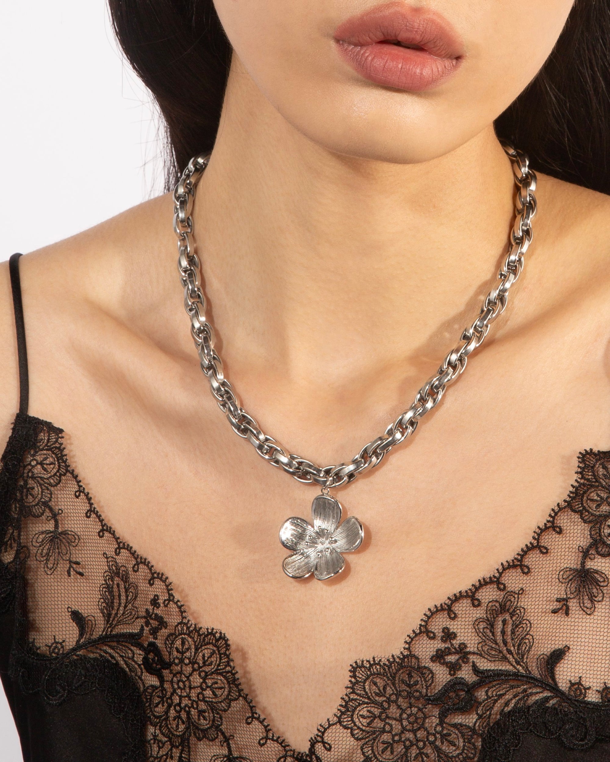 A person wearing a black lace top showcases the Blossom Necklace by For Art's Sake®, featuring a chunky silver chain with a large, detailed flower pendant, measuring 46cm in length.