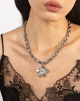 A person wearing a black lace top showcases the Blossom Necklace by For Art's Sake®, featuring a chunky silver chain with a large, detailed flower pendant, measuring 46cm in length.
