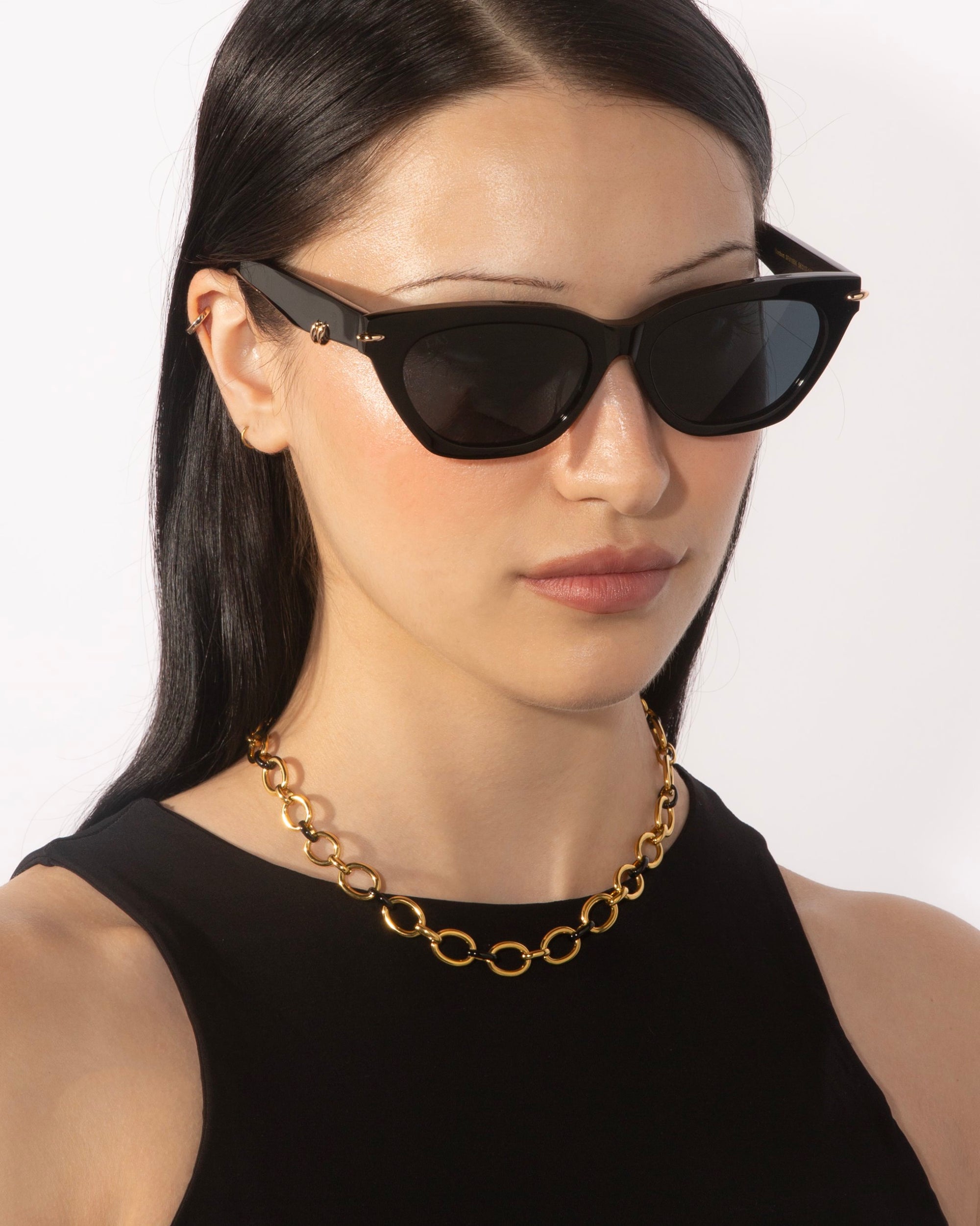 A person with long, dark hair wears cat-eye sunglasses and a black top, complemented by the bold Raya Necklace from the For Art's Sake® Love Letters collection. The background is a plain white wall.