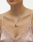 A woman showcases The Puffy Heart Pendant by For Art's Sake®—a gold puffed heart on a delicate chain—while wearing a pale pink lace camisole, set against a plain light background.