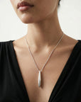 A woman models The Droplets Pendant Necklace by For Art's Sake®, featuring a sleek silver droplet pendant over a black top, with focus on her neck and collarbone against a plain, light background.