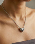 A person wears For Art's Sake® The Sculpted Heart Necklace—a shiny, heart-shaped pendant. The close-up highlights the necklace against bare skin above a strapless gray top.