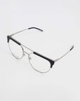 A pair of stylish Frosty prescription glasses by For Art's Sake® with a metallic frame and black accents on the top of the lenses, resting on a white surface. The design is minimalist with thin arms and clear lenses that include blue light filter technology.