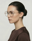 A woman with dark hair in a bun wears For Art's Sake® Dumpling oversized clear acetate frames with gold-plated arms and chain details, paired with a brown top, looking left against a light background.