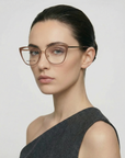 A woman with dark hair tied back wears For Art's Sake® Stella Two cat-eye glasses and a one-shoulder dark gray top, looking neutrally at the camera against a plain light background.