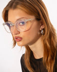 A person with long, light brown hair is wearing large-framed optical glasses in a cat-eye silhouette called Cadenza by For Art's Sake® and silver hoop earrings. They have a serious expression and are dressed in a black top, posing against a neutral background.