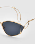 The Ornate sunglasses by For Art's Sake® feature gold frames, almond-shaped dark lenses, and slim decorative wire arms for a vintage look, shown against a plain light background.