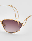 A close-up of For Art's Sake® Ornate sunglasses, featuring stylish gold almond-shaped frames, dark gradient lenses, and distinctive wire-detailed arms against a plain light background.