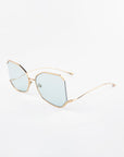 A pair of stylish eyeglasses with a geometric, hexagonal frame featuring light blue tinted lenses. The Painter by For Art's Sake® has a thin, gold-plated stainless steel frame and temples with a minimalist and modern design. The ultra-lightweight Nylon lenses offer 100% UVA & UVB protection. They are placed on a plain white background.