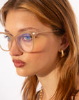A person with long, light brown hair is seen in a close-up side profile wearing For Art's Sake® Paris Two cat eye-inspired optics and gold hoop earrings. Their neutral expression contrasts with the plain white background.