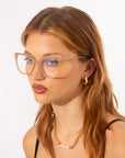 A person with long light brown hair wears For Art's Sake® Paris Two cat eye-inspired optics, gold hoop earrings, and a gold necklace with pearls. They have a neutral expression, wearing a black top against a plain white background.