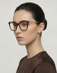 A woman with fair skin and dark hair in a low bun wears large brown Paris Two cat eye glasses by For Art's Sake® and a dark brown top, looking to the left against a plain light background.