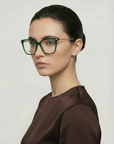 A woman with fair skin and dark hair in a neat updo wears For Art's Sake® Paris Two large green acetate eyewear and a brown top, looking slightly to the side against a plain, light background.