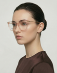 A woman with fair skin and dark hair in a low bun wears For Art's Sake® Paris Two—large, transparent acetate glasses—with a dark brown top, posing in profile against a plain, light background.