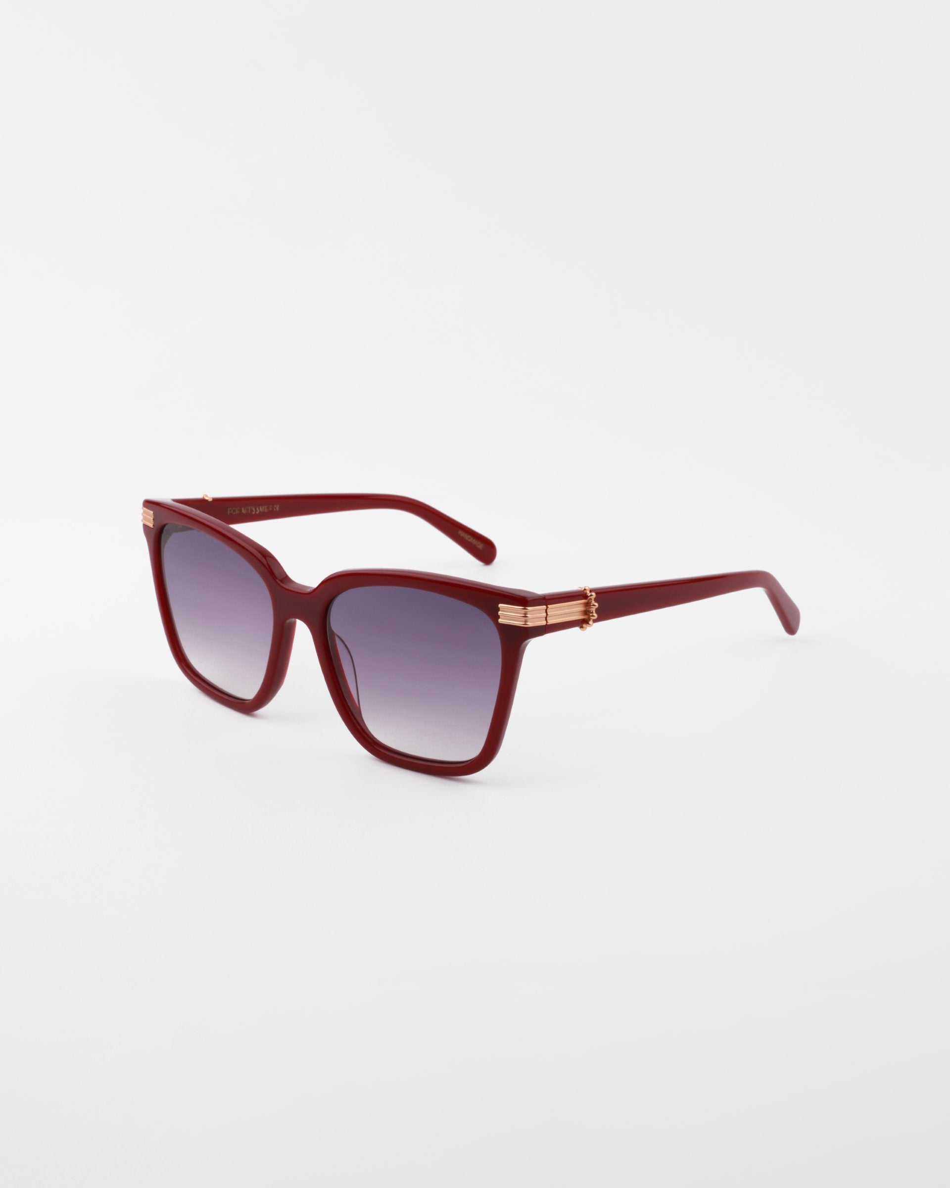 A pair of stylish, handmade acetate sunglasses featuring dark maroon frames and gradient purple, UV-protected lenses. The temples boast an 18-karat gold-plated decorative accent near the hinges. The Gaia sunglasses by For Art's Sake® are positioned on a white background.