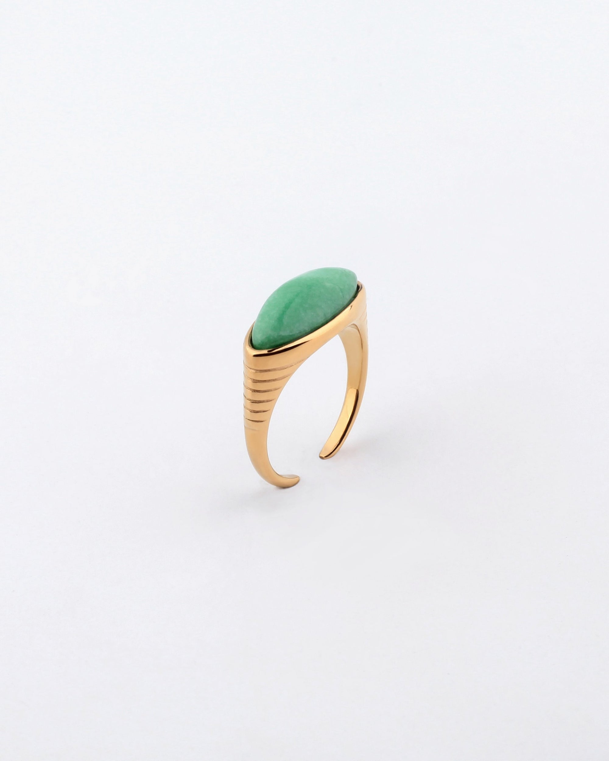 The Enza Ring by For Art's Sake® is a gold-plated stainless steel ring adorned with an elongated oval green gemstone on top, shown against a plain white background. It showcases a sleek, modern design with grooved detailing on the band and a polished enamel finish for added elegance.