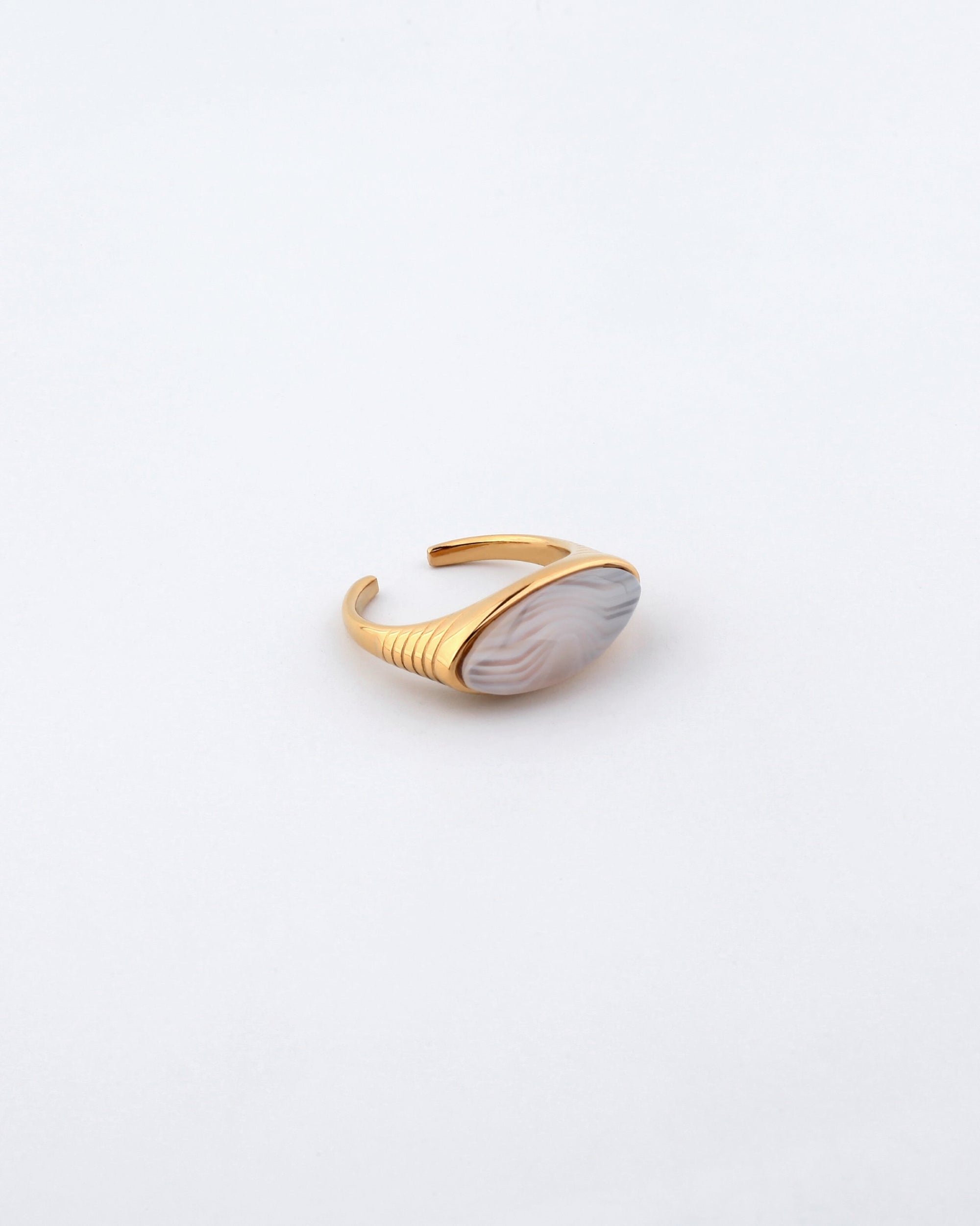The Enza Ring by For Art's Sake® is an elegant gold-plated stainless steel accessory, featuring an adjustable band. At its center sits an oval, marbled stone adorned with swirling hues of white and light grey, offering a unique, natural aesthetic. The band showcases a sleek and minimalist design with a polished enamel finish and is slightly open at the back for easy fit adjustment.