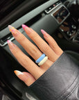 A hand with vibrant nail art showcasing a butterfly design on the ring finger is elegantly adorned with a minimalist Raya Ring by For Art's Sake®. The individual wears a black leather jacket accompanied by two chunky rings in shades of blue and cream. The backdrop displays the sleek interior of a car.