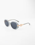 A pair of stylish Seaside sunglasses by For Art's Sake® featuring round black lenses with a light gray, semi-translucent, handmade acetate frame. The temples have gold-plated detailing near the hinges, adding a touch of elegance to the design. The background is plain white.