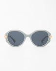 A pair of light gray oval-shaped Seaside sunglasses by For Art's Sake® with dark blue, shatter-resistant lenses. The handmade acetate frame features gold-plated temple detailing on a plain white background.