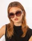 A person with light brown, shoulder-length hair is wearing large, Helios sunglasses by For Art's Sake® with a brown tint. They have gold hoop earrings and are dressed in a sleeveless black top. The luxury eyewear adds a touch of sophistication against the plain white background.