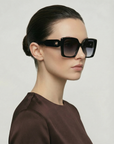A woman with dark hair in a low bun wears For Art's Sake® Eos oversized soft-square sunglasses and a brown top, posing against a plain light background.