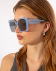 A woman with long, light brown hair wears oversized Eos - Blue sunglasses by For Art's Sake®, pairing them with small, dangling earrings and a sleeveless gray top. Only 2 left of these soft-square frames. Elegance stands out against the plain background.
