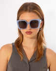 A woman with light brown hair wears For Art's Sake® Eos - Blue oversized, soft-square sunglasses (ONLY 2 LEFT) and a sleeveless gray top, looking at the camera against a plain white background.